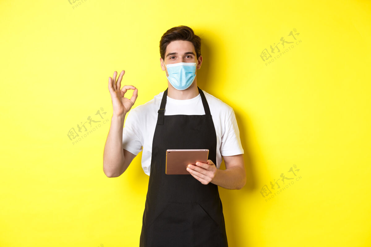 面膜covid-19的概念 小型企业和流行病戴着医用面罩和黑色围裙的销售员显示ok标志 用黄色背景的数字平板电脑接受订单男人黄色时尚