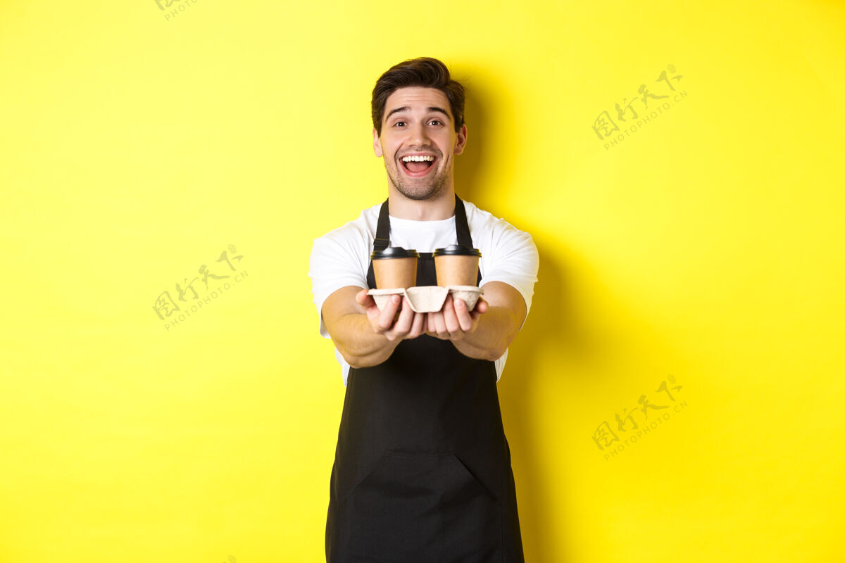 年轻友好的咖啡师 穿着黑色围裙 端着两杯咖啡 微笑着 站在黄色的背景上展台咖啡师杯子