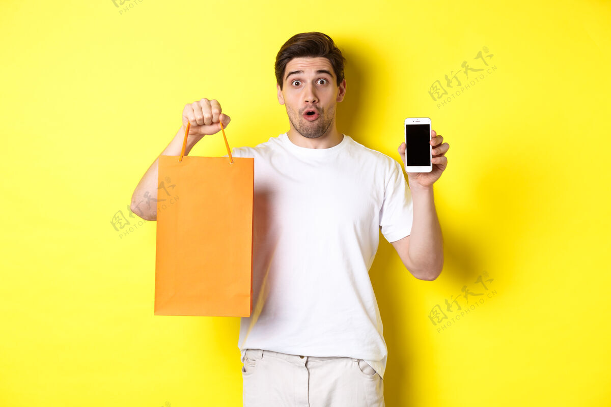 站着惊讶的男人展示手机屏幕和购物袋 站在黄色背景下复制空间工作室男人表情