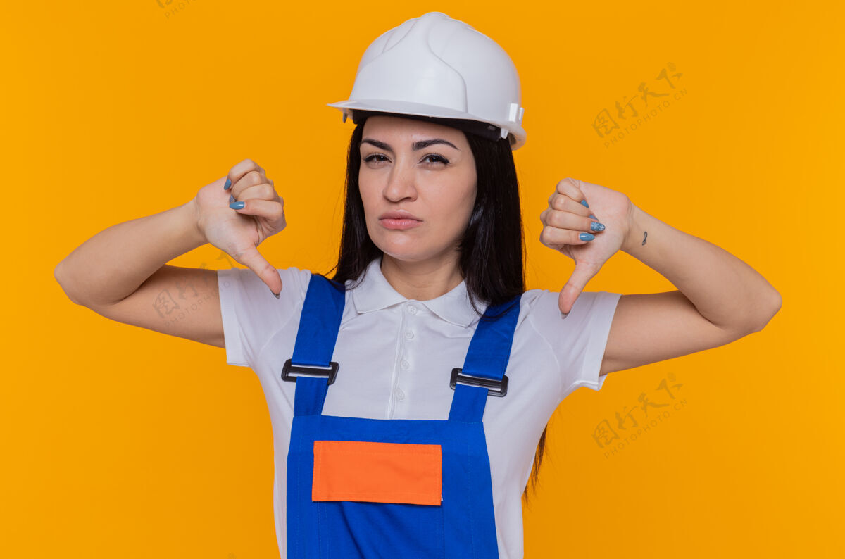 倒下身穿建筑制服 头戴安全帽的年轻建筑工人站在橙色的墙上 不高兴地竖起大拇指看着前面安全制服女人