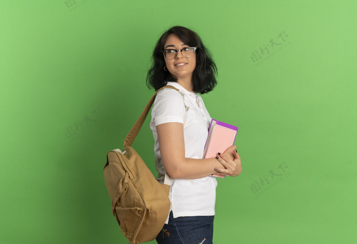 包年轻漂亮的高加索女生戴着眼镜 背着书包站在一边 手里拿着书 在绿色的草地上回望 留着复印空间女学生背请