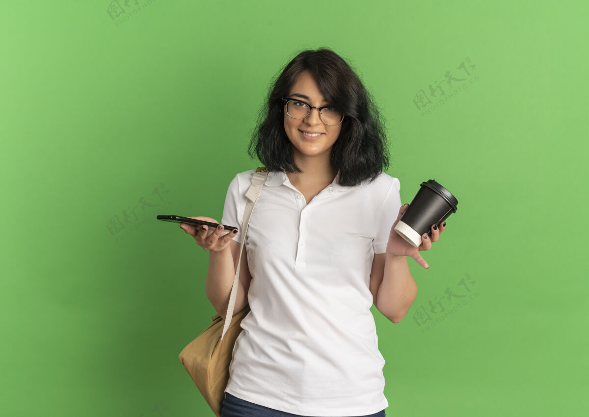 背部年轻的高加索女孩 面带微笑 戴着眼镜 背着书包 拿着电话和咖啡杯 放在绿色的空间里眼镜咖啡复制