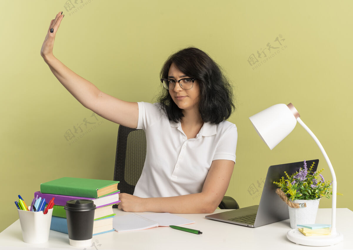漂亮年轻自信漂亮的白人女学生戴着眼镜高举着手坐在书桌旁 绿色的桌子上放着学习用具 还有复印空间自信穿绿色