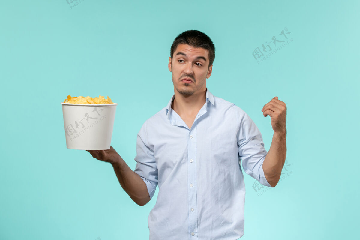 容器正面图：年轻的男性手拿篮子 浅蓝色的表面上放着薯片远程男性杯子