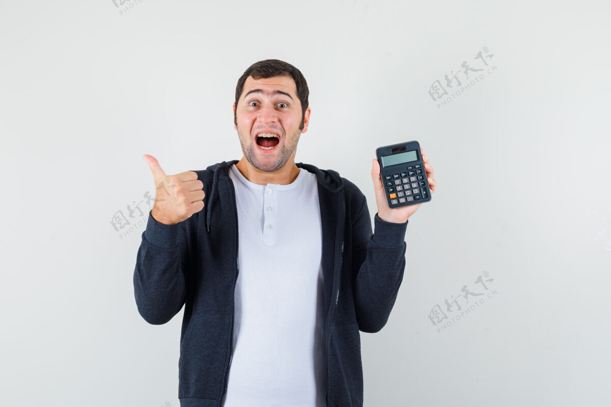 年轻年轻人在白色t恤和拉链前黑色连帽衫举行计算器 并显示拇指向上 看起来乐观 正面视图一男性前面