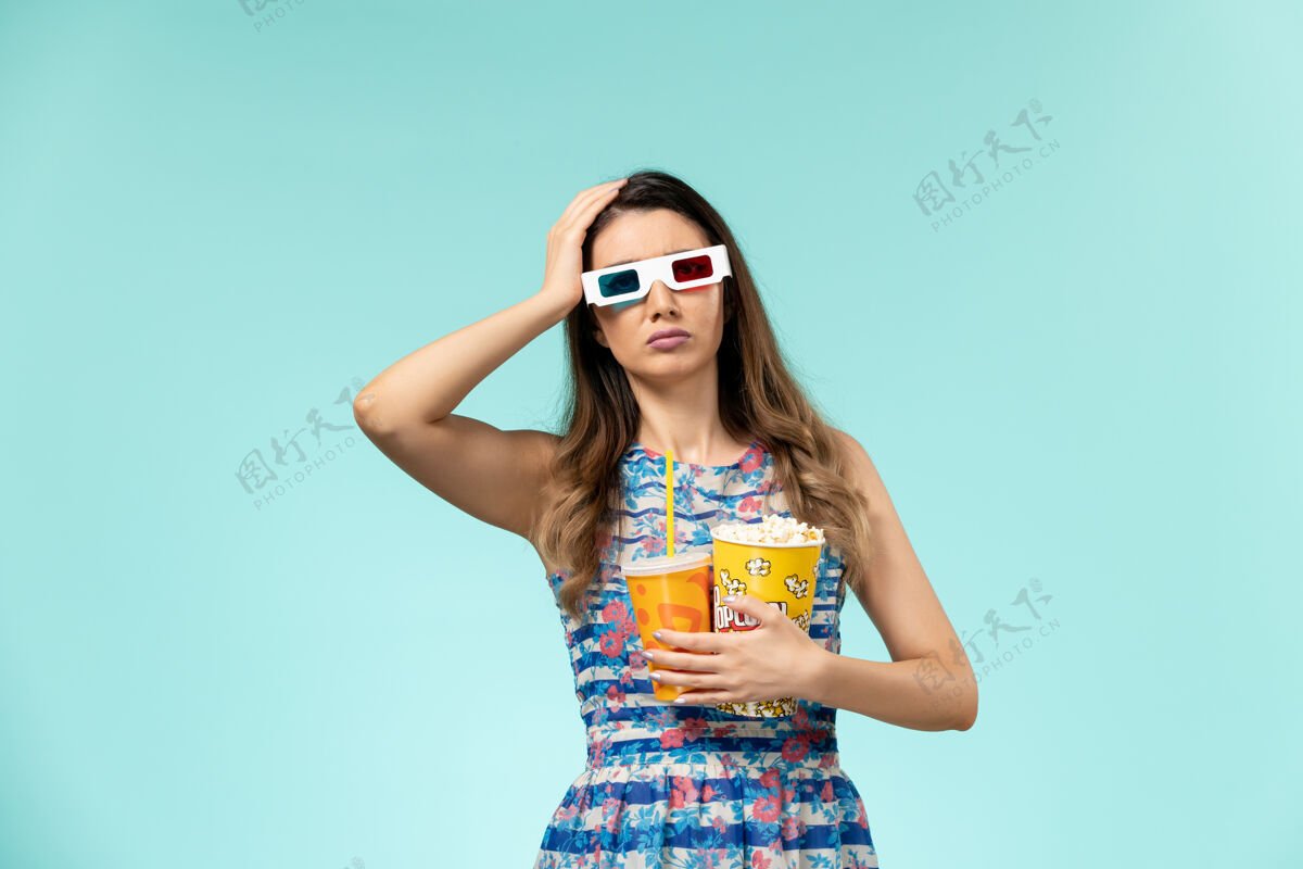 性感正面图年轻女性 带着爆米花包 戴着d墨镜在蓝色表面喝酒正面年轻女性电影院