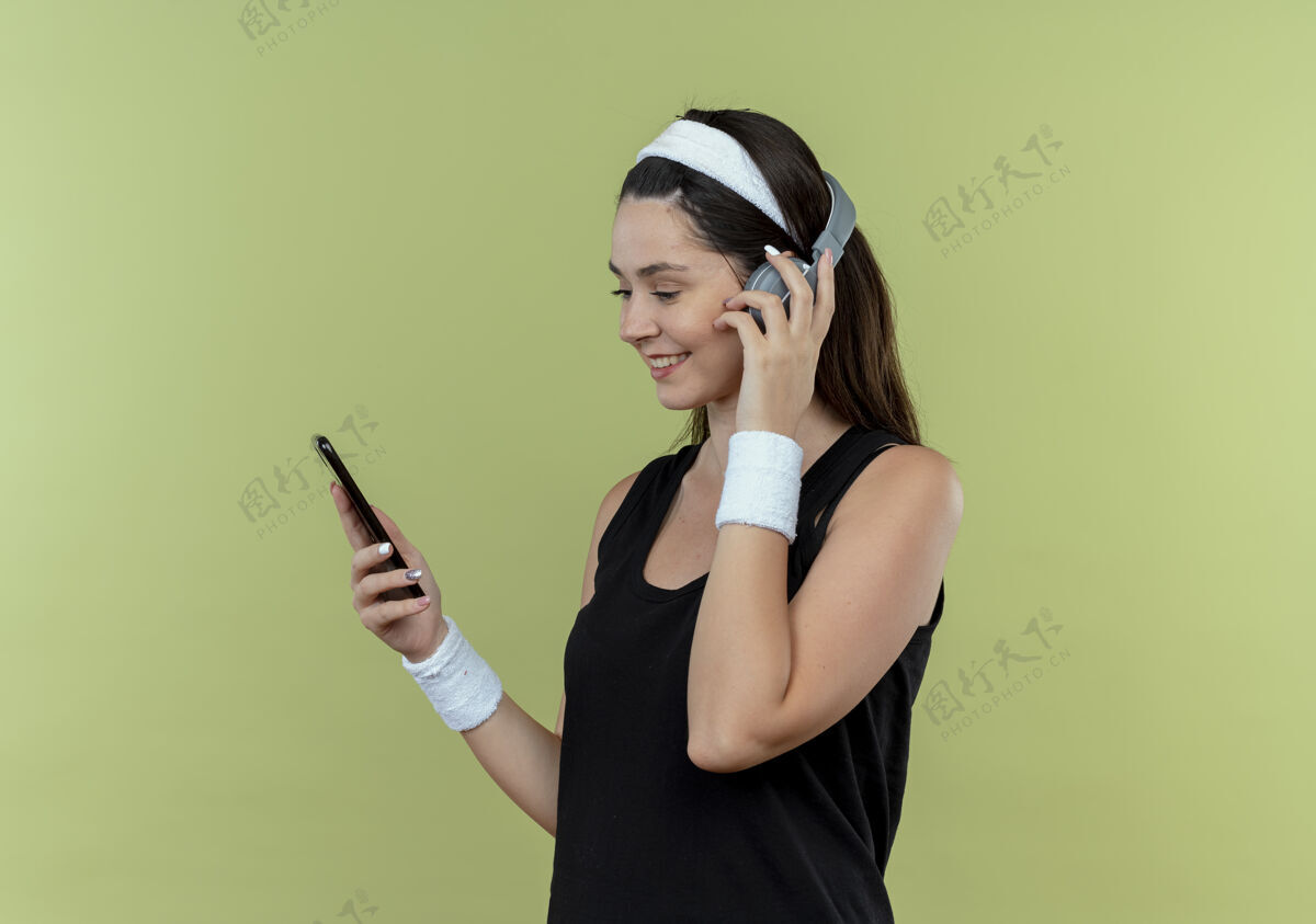 年轻戴着耳机戴着头带的年轻健身女士站在灯光墙的上方看着智能手机的屏幕搜索音乐屏幕音乐灯光