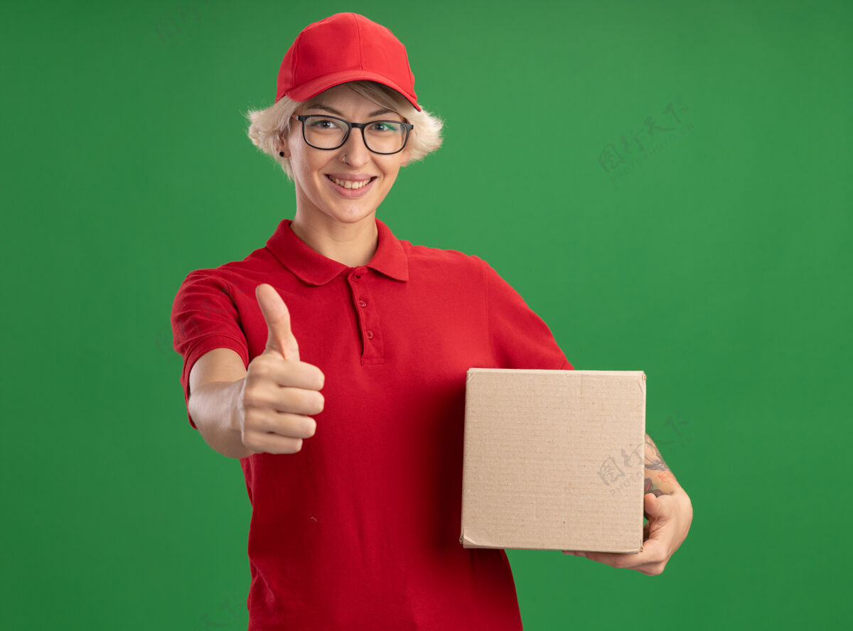 抱着年轻的送货员 身穿红色制服 戴着帽子 戴着眼镜 拿着纸板箱 站在绿色的墙边兴高采烈地微笑着 竖起大拇指戴着欢呼展示