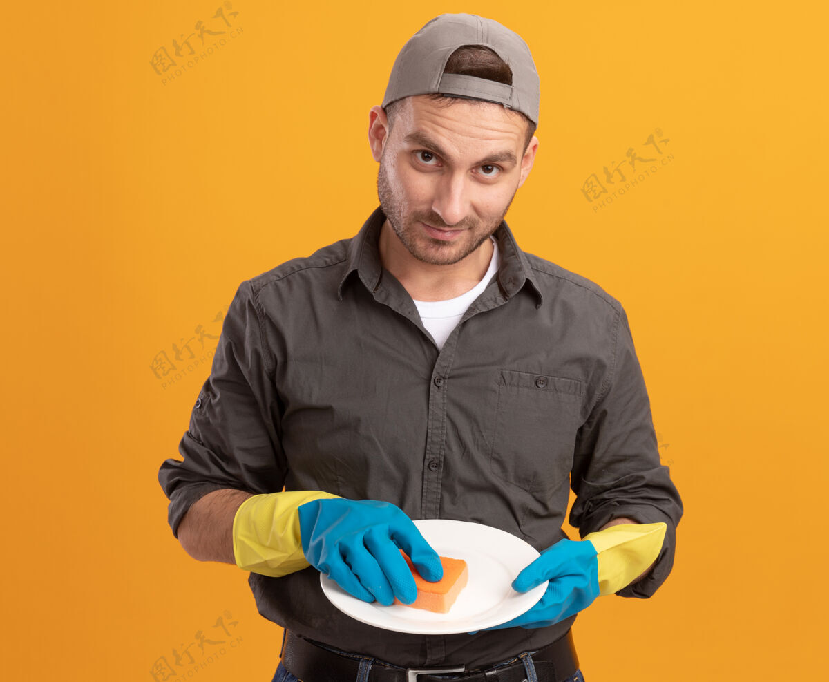 洗年轻的保洁员穿着休闲服 戴着橡胶手套 拿着盘子和海绵 站在橘色的墙上 满脸笑容 自信满满盘子手套自信