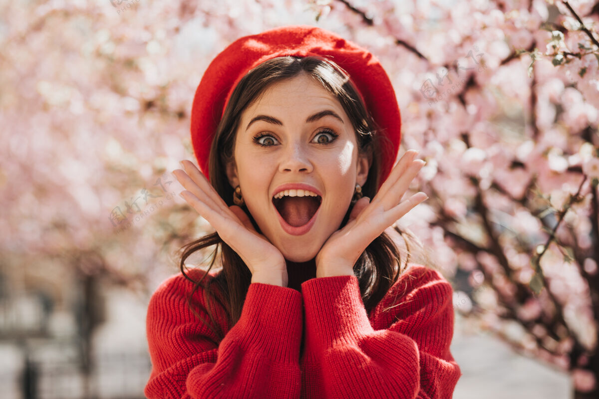 春天在樱花的背景下 戴着红色贝雷帽的女孩欣喜若狂地看着镜头在盛开的花园里 穿着毛衣的绿眼睛女人惊讶地摆出姿势自行车生活连衣裙