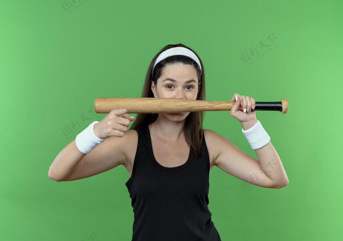指着戴着头巾的年轻健身女士手持棒球棒 手指微笑着站在绿色的墙上微笑女人棒球
