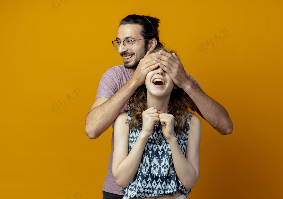 微笑年轻的夫妇男人闭上女孩的眼睛 而她微笑着高兴和兴奋的橙色墙年轻兴奋夫妇