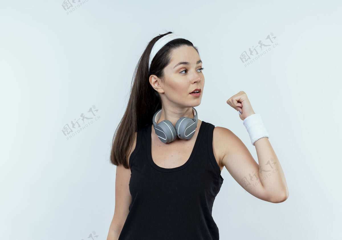 耳机戴着耳机戴着头带的年轻健身女士站在白墙上 握紧拳头向一旁望去头带站立女人