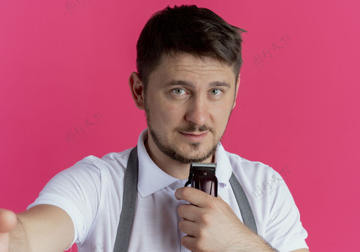 围裙围裙上的理发师拿着剃须刀 脸上带着微笑站在粉红色的墙上男人修剪站