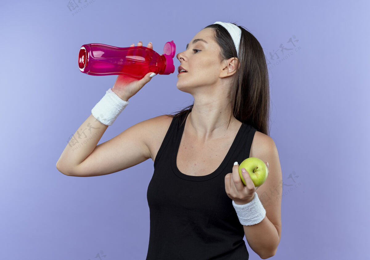 喝酒戴着头巾的年轻健身女士 在锻炼后拿着绿色苹果饮用水站在蓝色的墙上站着抱着年轻