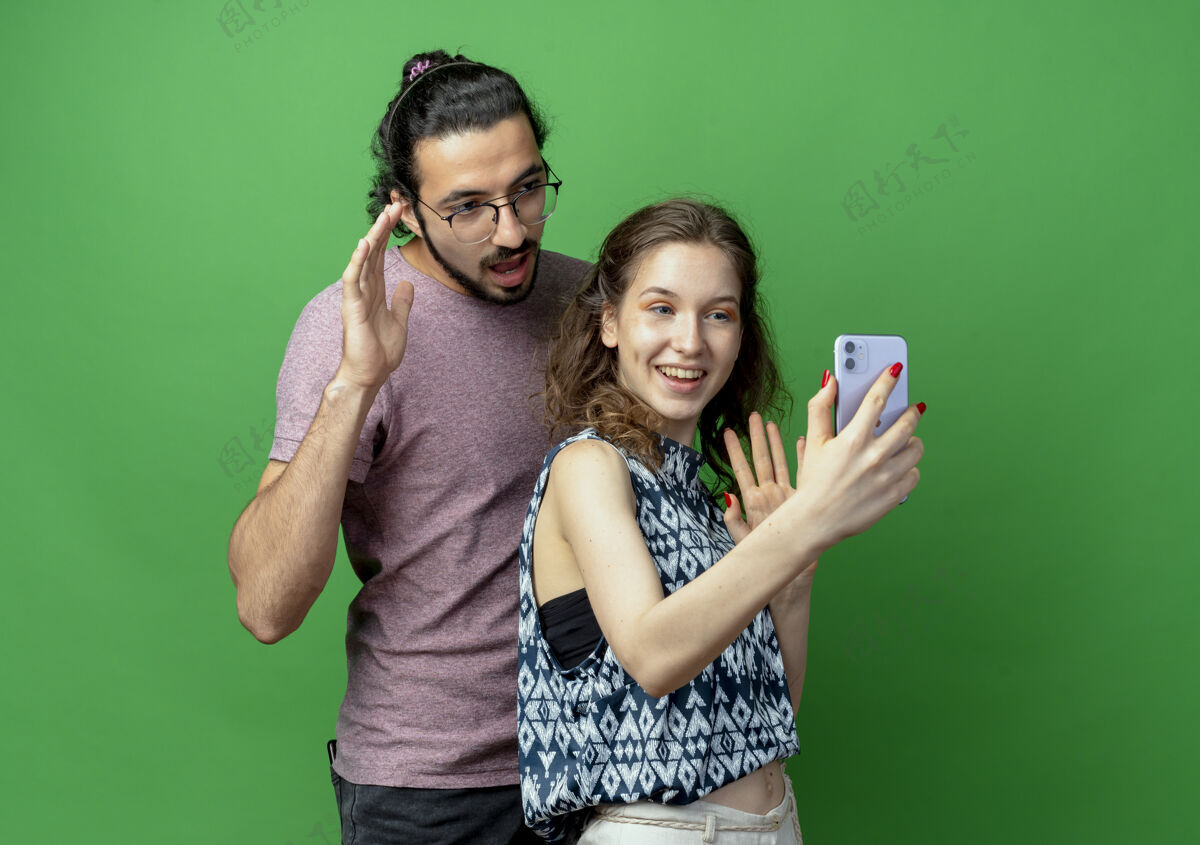 用着一对年轻的男女 幸福的女人用智能手机拍下他们站在绿色的墙上站着年轻人拿着