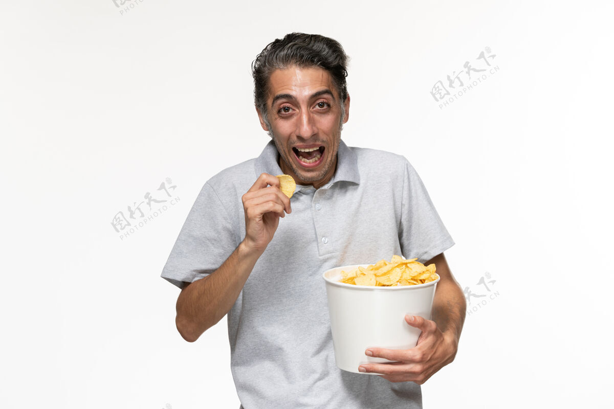 笑着正面图年轻的男性吃着薯片 在浅白的表面上欢笑男果汁早餐