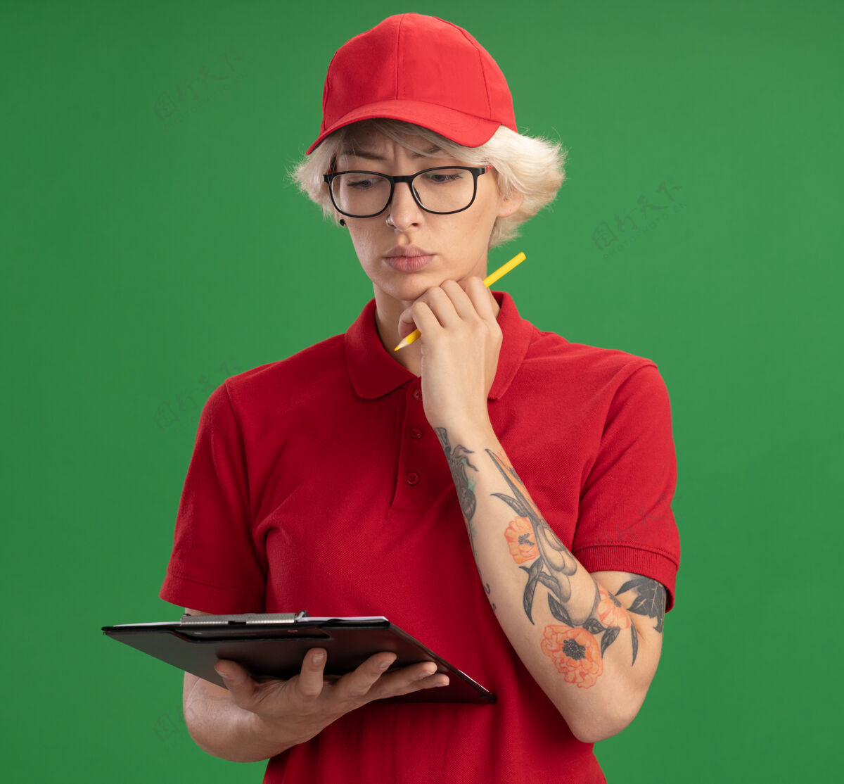 穿身穿红色制服 戴着帽子的年轻送货员戴着一副眼镜 上面有空白页和铅笔的剪贴板 站在绿色的墙壁上 面带严肃的思考站眼镜交货