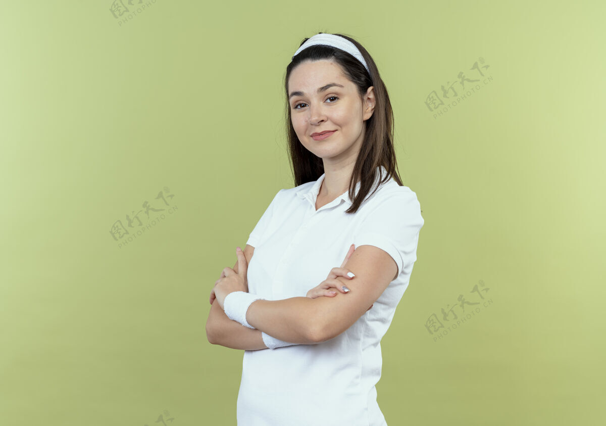 年轻戴着头巾 双臂交叉 表情自信的年轻健身女士站在轻盈的墙壁上站立健身头带