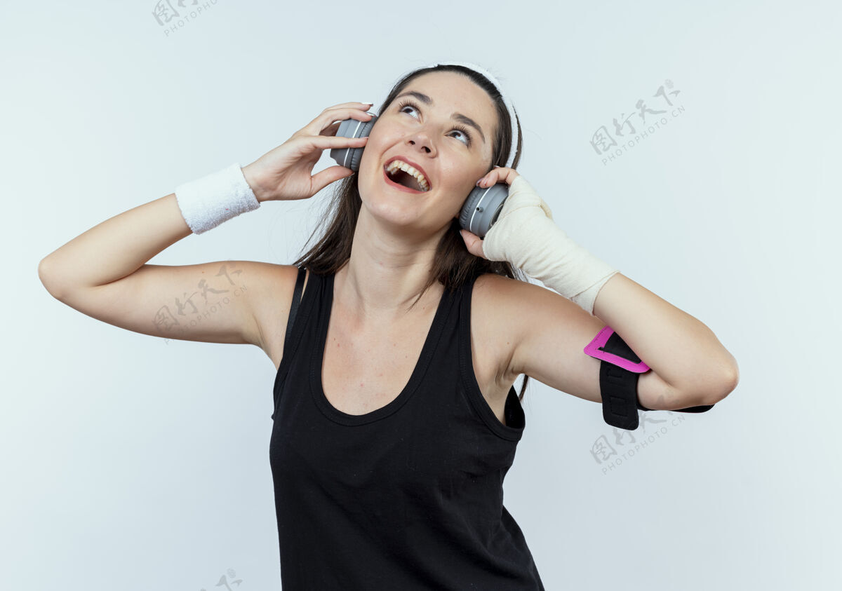 享受戴着耳机和智能手机臂章的年轻健身女士站在白墙上欣赏着她最喜欢的音乐音乐年轻健身