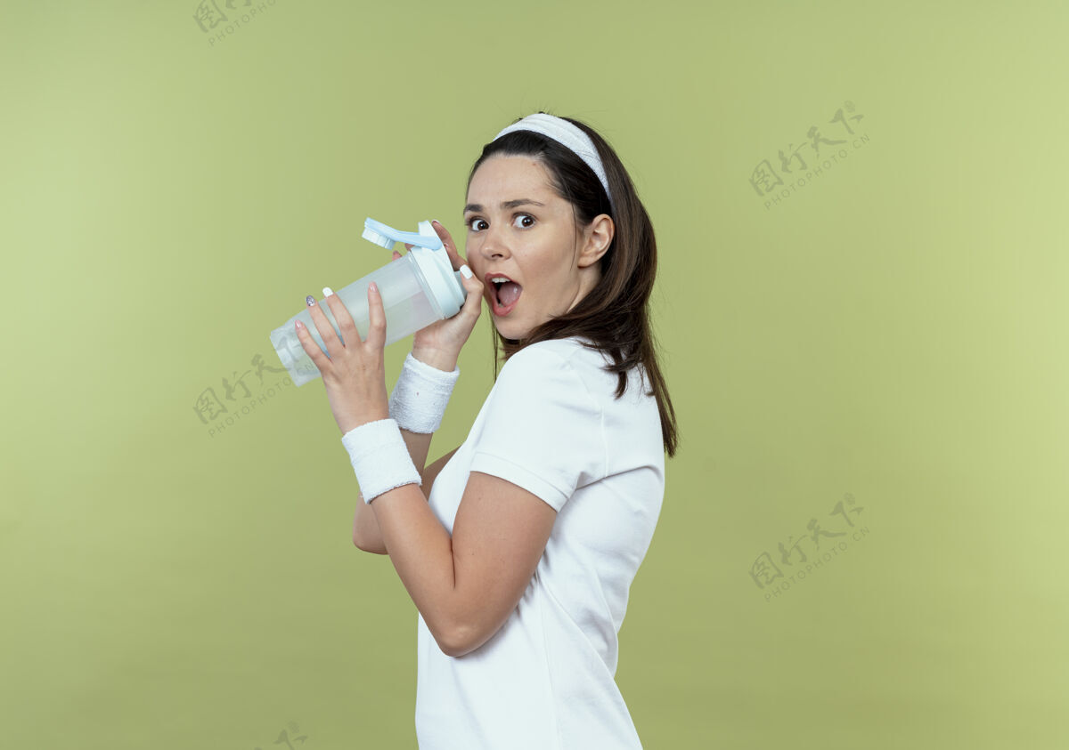 瓶子戴着头巾拿着一瓶水的年轻健身女士站在轻薄的墙上大吃一惊女人站着年轻