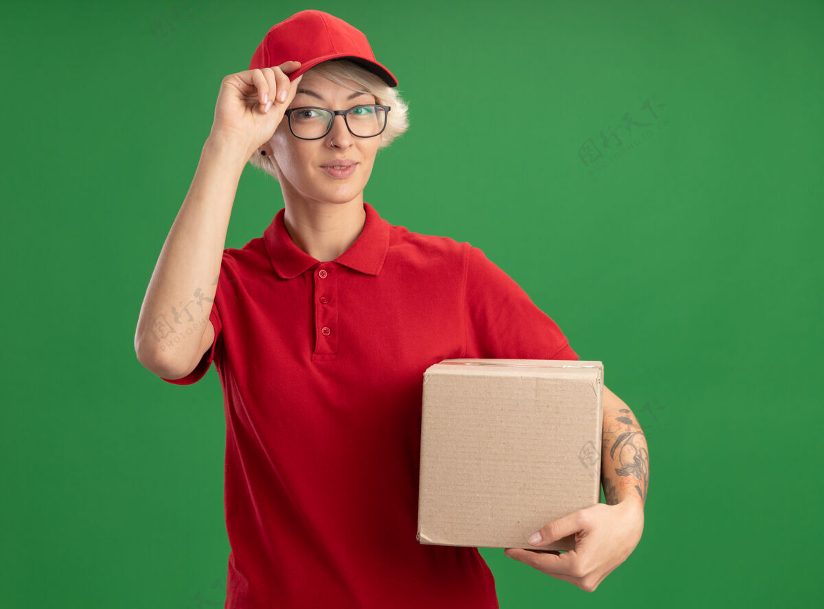 帽子年轻的送货员身穿红色制服 戴着帽子 戴着眼镜 手里拿着纸板箱 神情自信地抚摸着站在绿色墙上的帽子立场眼镜举行