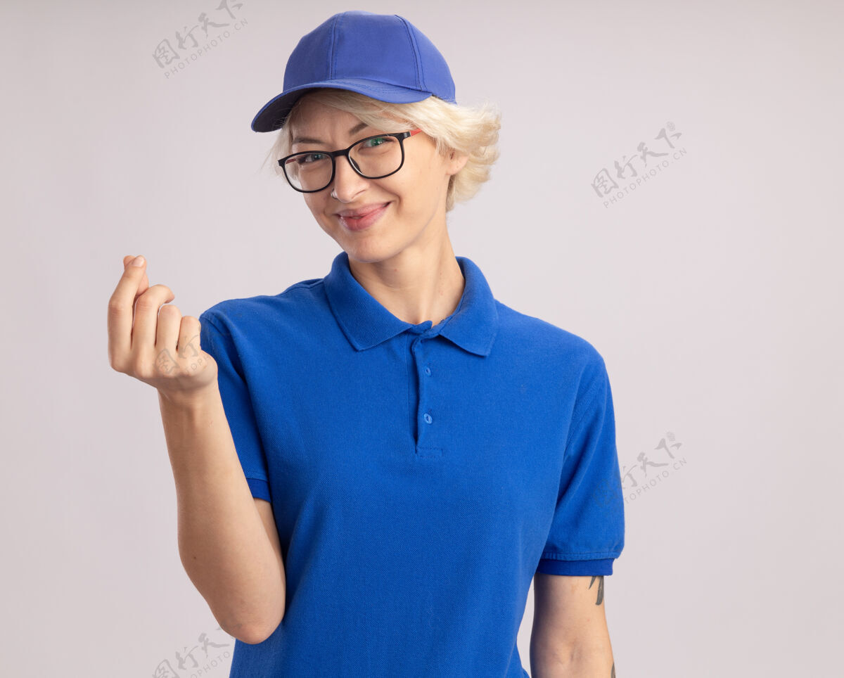 手势身穿蓝色制服 戴着眼镜 面带微笑的年轻送货员站在白色的墙上 用手指搓着赚钱的手势擦穿钱