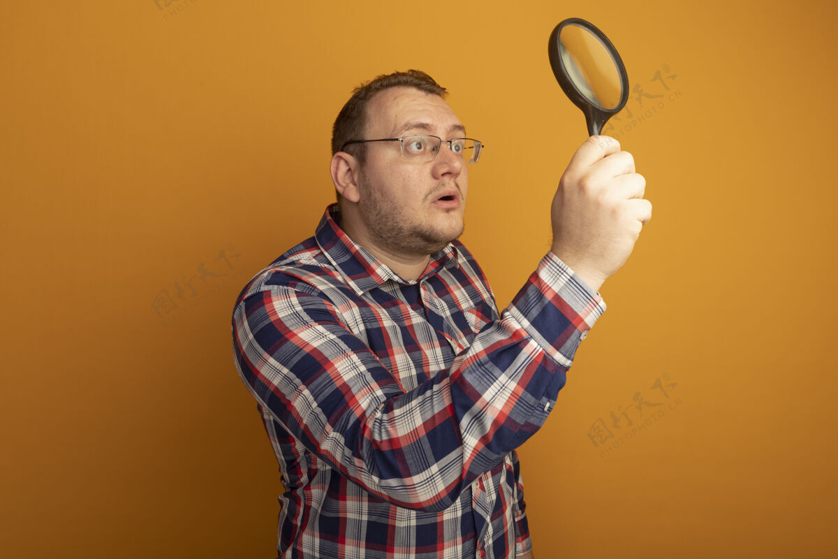 男人一个戴着眼镜和格子衬衫的男人拿着放大镜站在橘色的墙上看着它很好奇站着眼镜放大镜