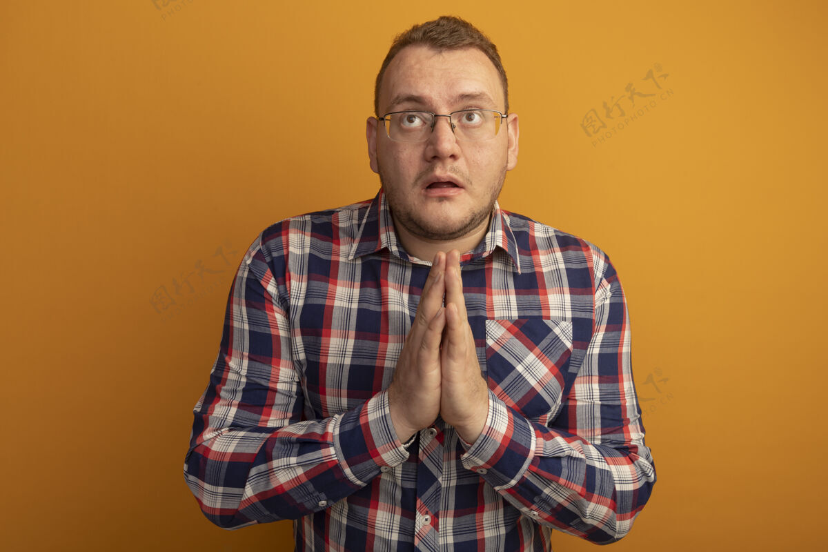 在一起戴着眼镜和格子衬衫的男人手拉着手 像是在祈祷 带着希望的表情站在橙色的墙上检查抱着表情