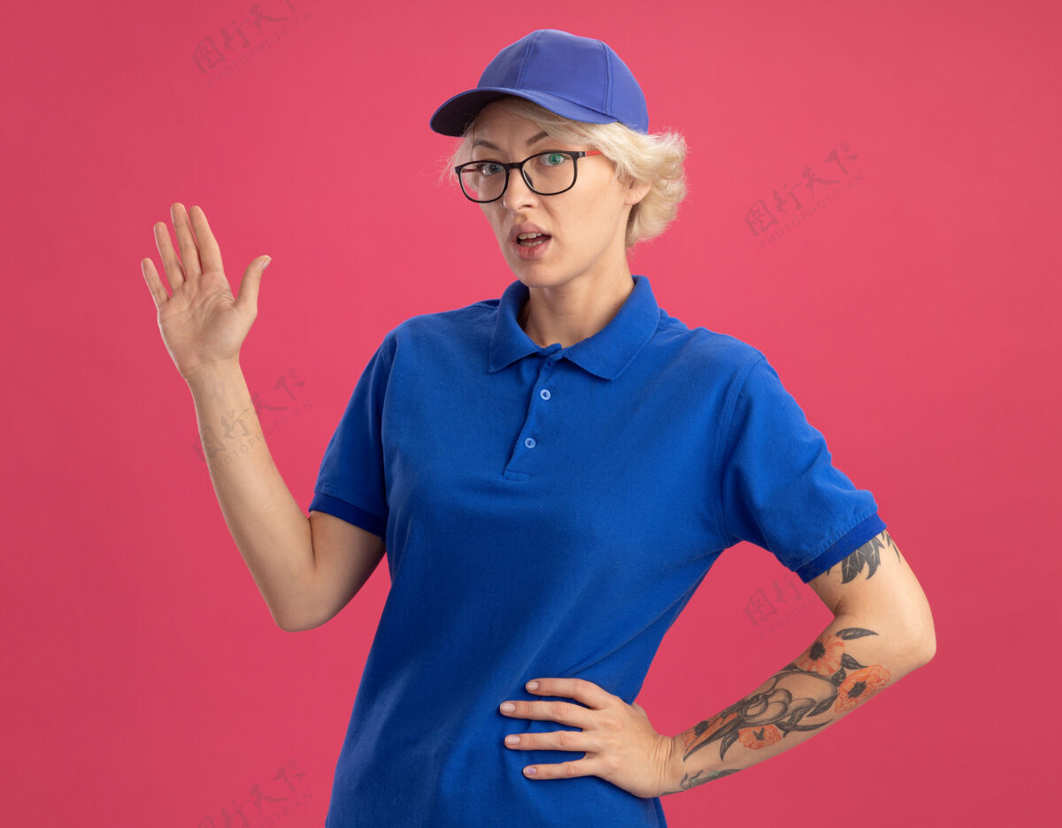 交货送外卖的女人穿着蓝色制服 戴着帽子 看起来很困惑 胳膊哦 手放在粉红色的墙上女人手臂制服