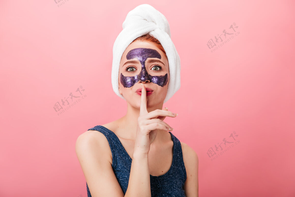 秘密美丽的女人在做水疗时展示秘密标志的前视图粉红色背景上隔离面具的漂亮女孩的摄影棚照片女性面膜治疗