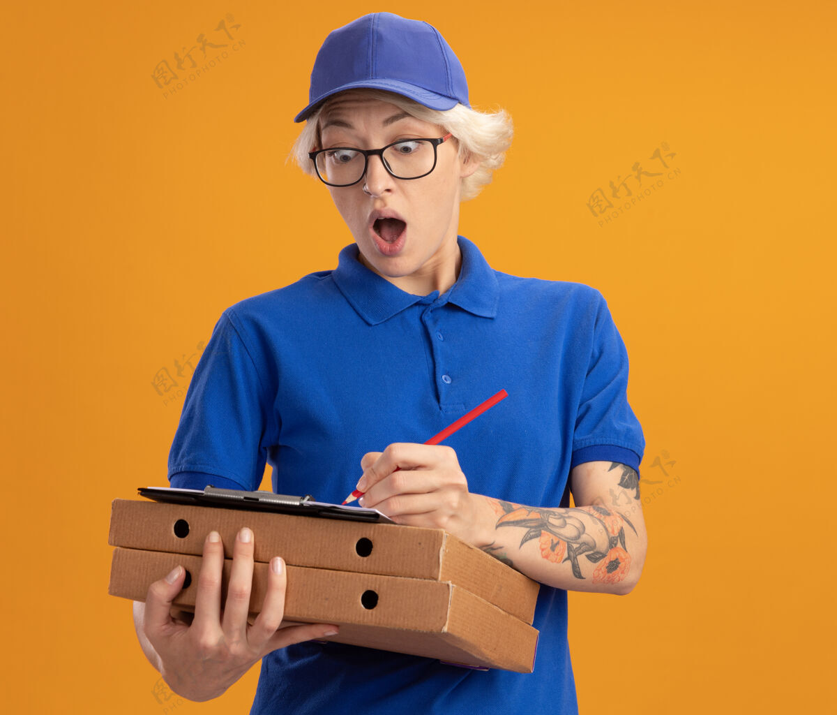 女人一个穿着蓝色制服 戴着眼镜 拿着披萨盒 拿着空白页的剪贴板 用铅笔在橘色墙上写东西的年轻送货员惊讶不已写字年轻盒子