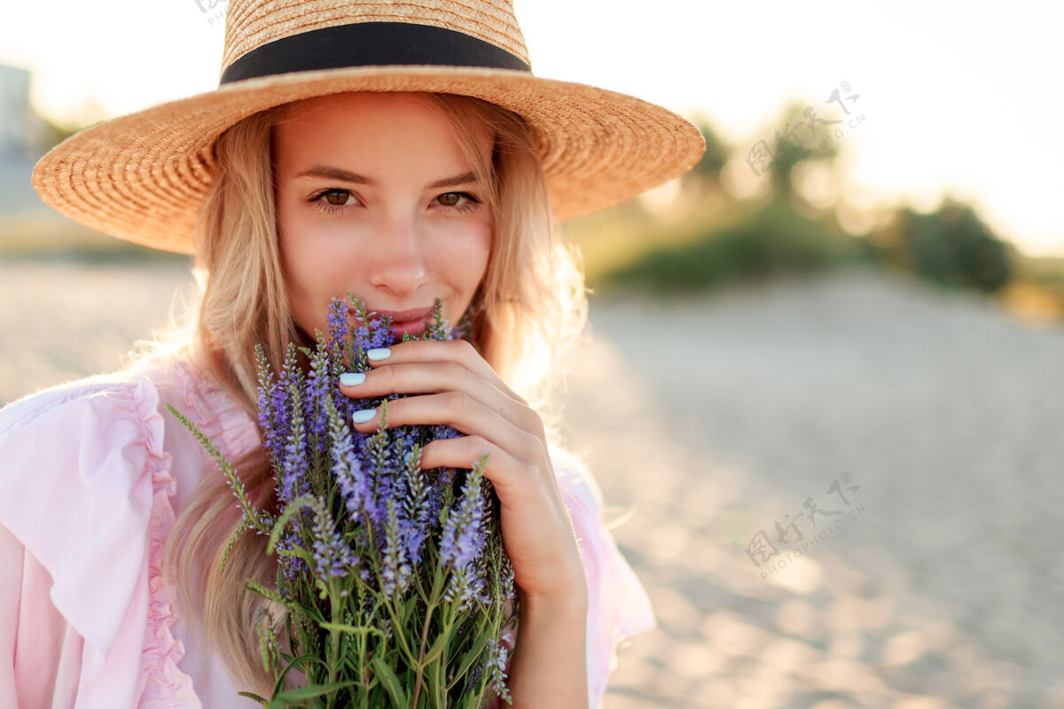 成人微笑着戴草帽的漂亮女孩在海边阳光明媚的海滩上摆着一束鲜花特写肖像女性花微笑