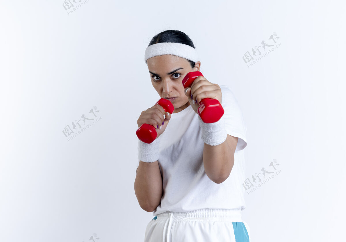 防守戴着头带和腕带的年轻自信的白种人运动型女人假装在白色空间里单独拿着哑铃进行辩护头带年轻腕带