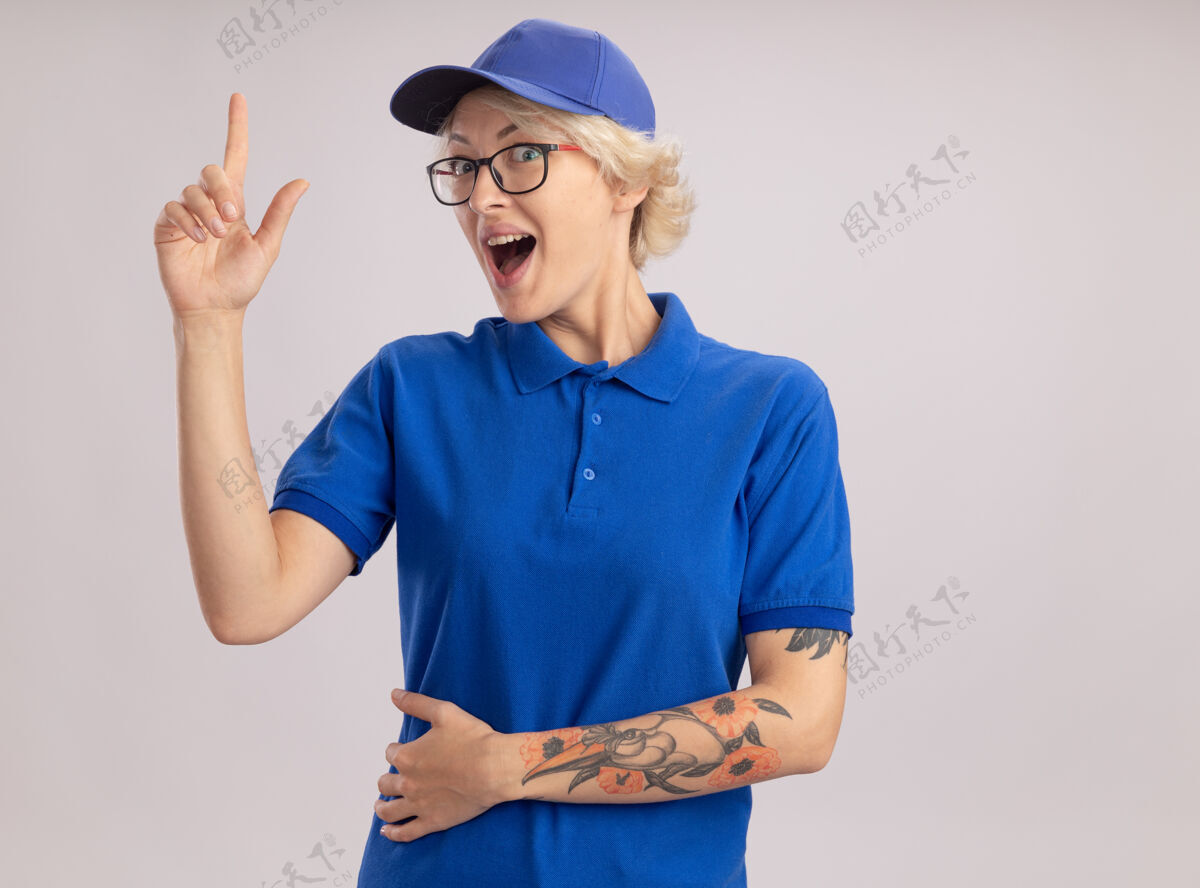 手指身穿蓝色制服 头戴鸭舌帽的年轻女送货员高兴地伸出食指站在白色的墙上交货欢呼女人