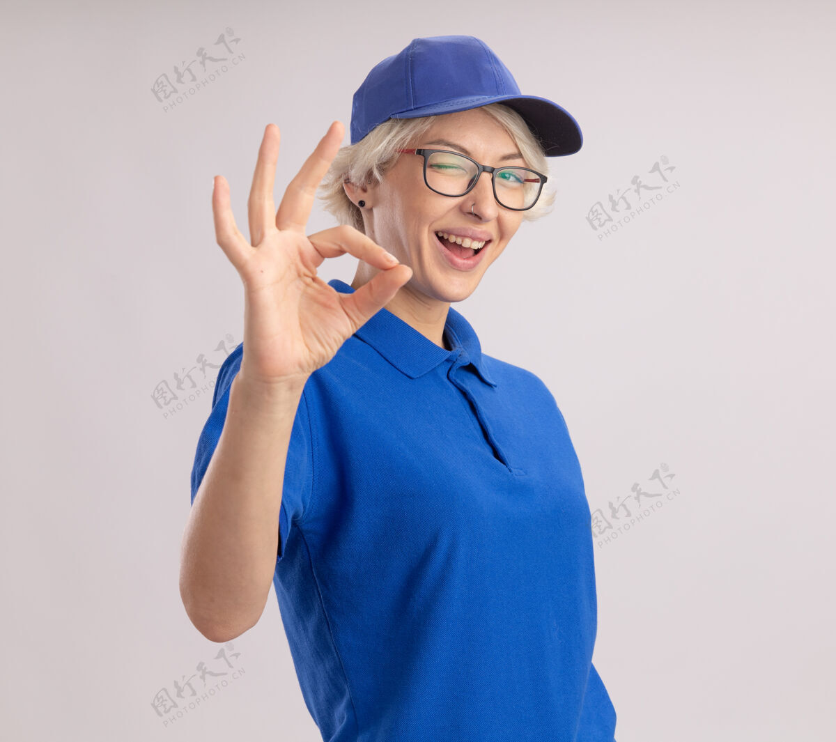 展示身穿蓝色制服 戴着帽子的年轻送货员 面带微笑 眨眼着 白墙上挂着“ok”的牌子眨眼站着女人