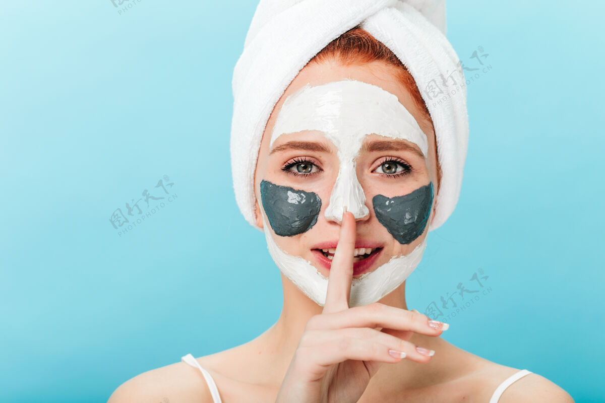 治疗戴着面具的令人毛骨悚然的女士的正面图显示了沉默的标志放松的女模特头上戴着毛巾在蓝色背景下的摄影棚拍摄皮肤健康清洁面部
