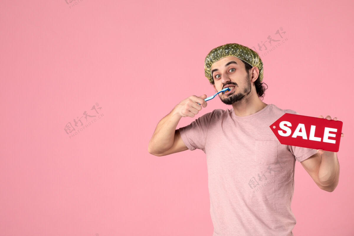 视图正面图年轻的男性正在清洁他的牙齿 拿着粉红色背景上的销售铭牌持有牙齿美容