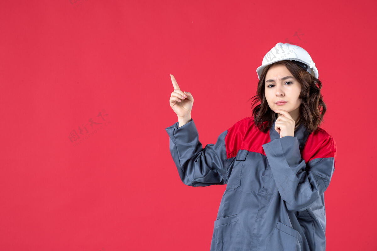 吸引力正面近距离观看穿着制服 戴着安全帽 指着孤立的红墙的思考型女建筑工人微笑成人制服