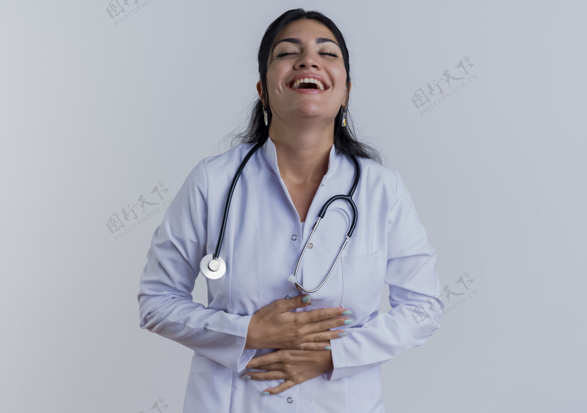 穿笑着的年轻女医生穿着医用长袍 手持听诊器 紧闭着眼睛 在白色的墙壁上隔离着复印空间年轻听诊器医疗