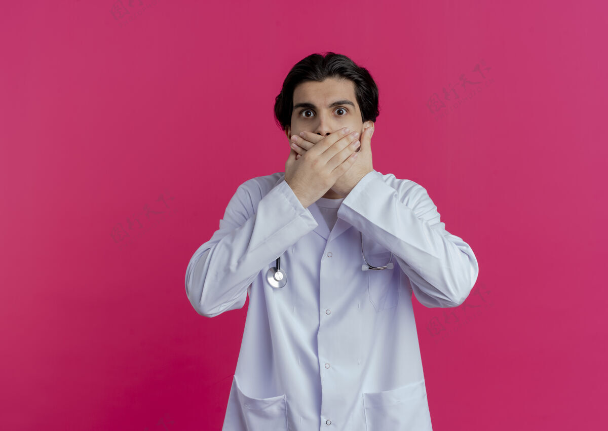 男性穿着医用长袍和听诊器的年轻男医生惊讶地将手放在隔离的粉红色墙上 并留有复印空间期待年轻长袍