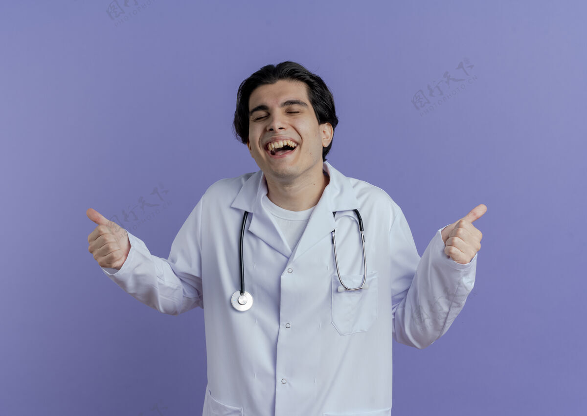 长袍快乐的年轻男医生穿着医用长袍 听诊器竖起大拇指 闭着眼睛 隔离在紫色的墙上表演穿医生