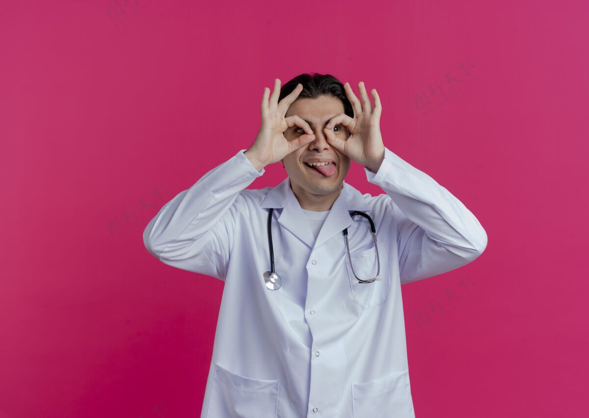 医生顽皮的年轻男医生 穿着医用长袍 戴着听诊器 一边看一边做着看的动作 用手做望远镜 把舌头孤立在粉红色的墙上 留着复印空间表演看年轻人