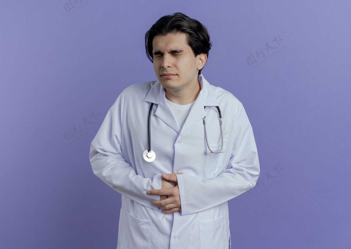 听诊器疼痛的年轻男医生穿着医用长袍 手持听诊器 紧闭着眼睛 在紫色的墙壁上隔离着 留着复印空间医生穿肚子