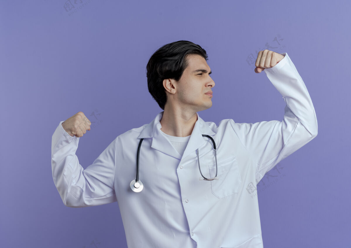 手势自信的年轻男医生穿着医用长袍 用听诊器做着有力的手势看着隔离在紫色墙上的拳头男性表情长袍