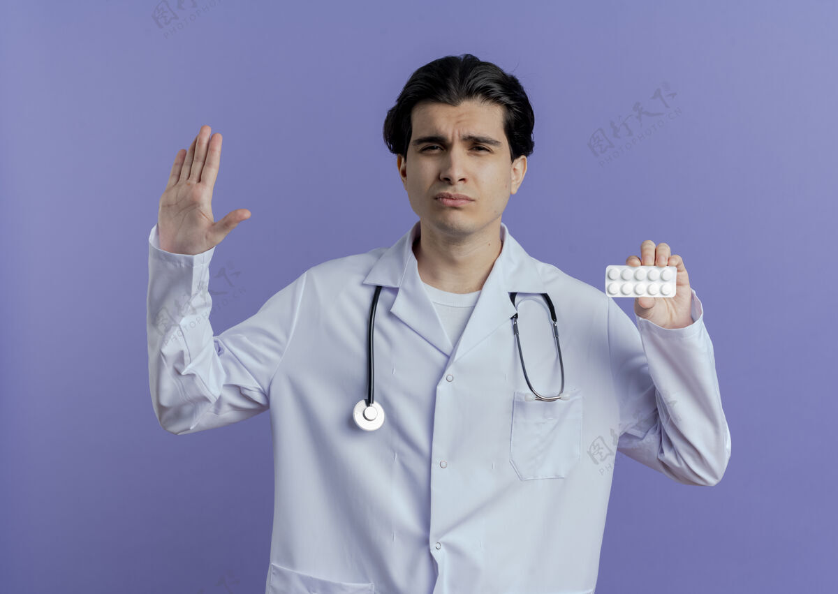 显示穿着医用长袍和听诊器的年轻男性医生 在紫色的墙上展示着一包药片在做停止动作医生不愉快手势