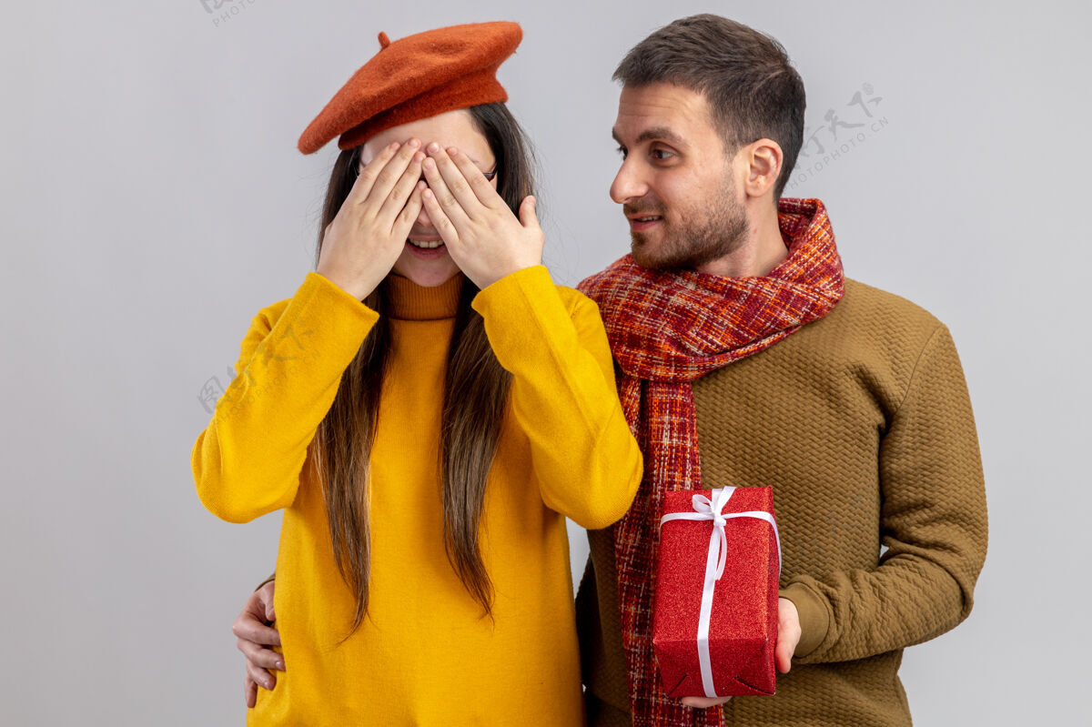 掩护年轻漂亮的情侣幸福的男人给他戴着贝雷帽微笑的女朋友送礼物 她双手捂着眼睛 一起欢庆情人节站在白色的背景下手给予白天