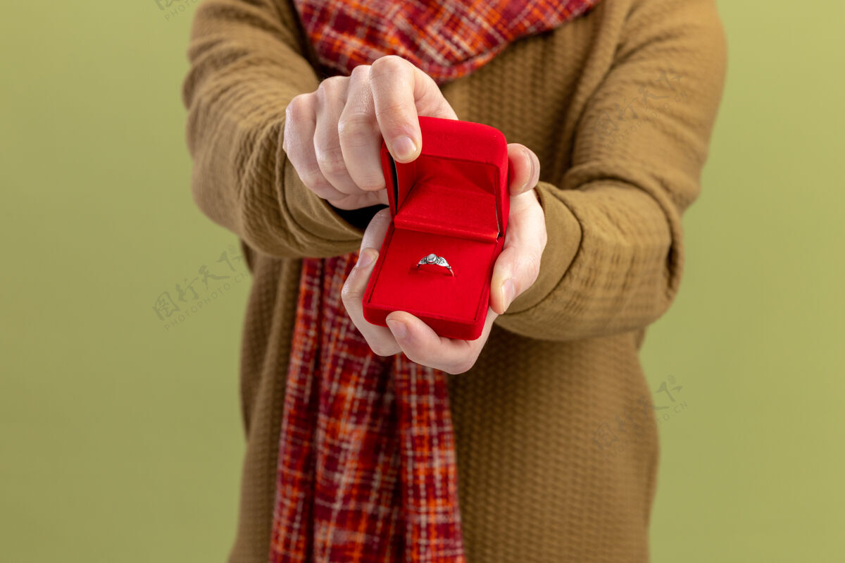 站身穿休闲服 脖子上围着围巾的年轻人在绿色的墙上展示着红色的盒子和订婚戒指情人节的概念戒指年轻男人