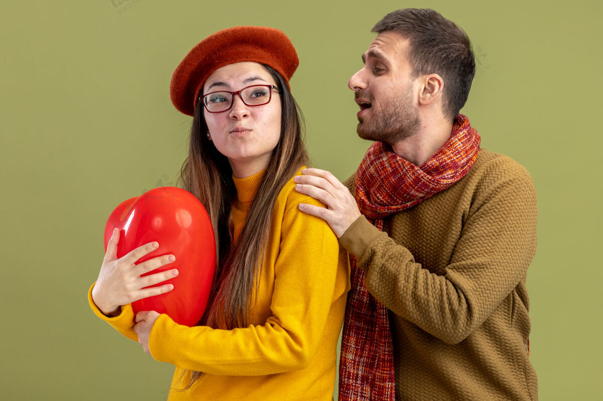 女朋友年轻漂亮的夫妇幸福的男人触摸他困惑的女友贝雷帽与心形气球庆祝情人节站在绿色的墙壁情侣日子贝雷帽