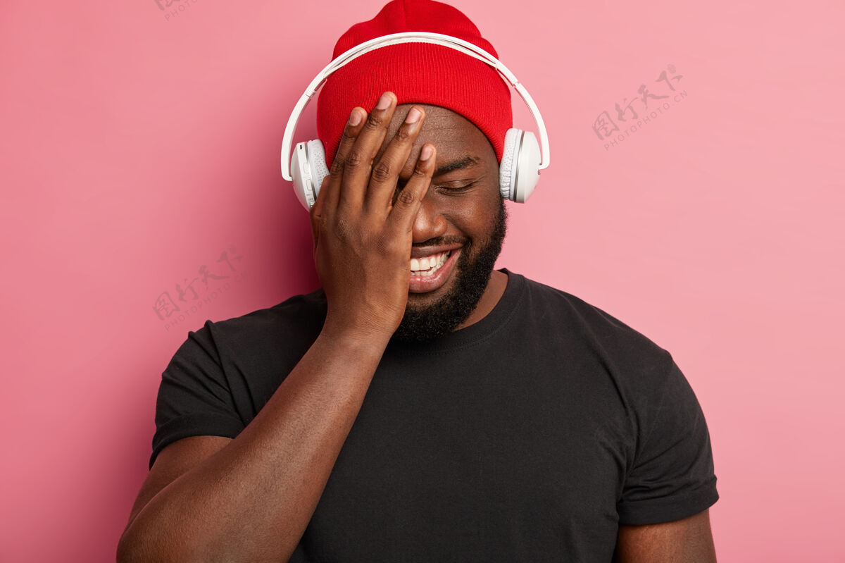 高兴戴着红帽的快乐的家伙 闭上眼睛 笑容灿烂 选择放松的歌曲戴着立体声耳机 隔离在粉红色的墙上 闲暇时间听音乐欢呼耳机爱好